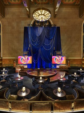 The Oscars 2021 set at the Union Station photographed from high ground. It's staged in an art-deco style, with curved booths with dark blue velvet upholstery, and round tables with lamps on them. The sitting arrangements are looking at the stage, which has a deep blue velvet curtain as a background with two screens on each side.