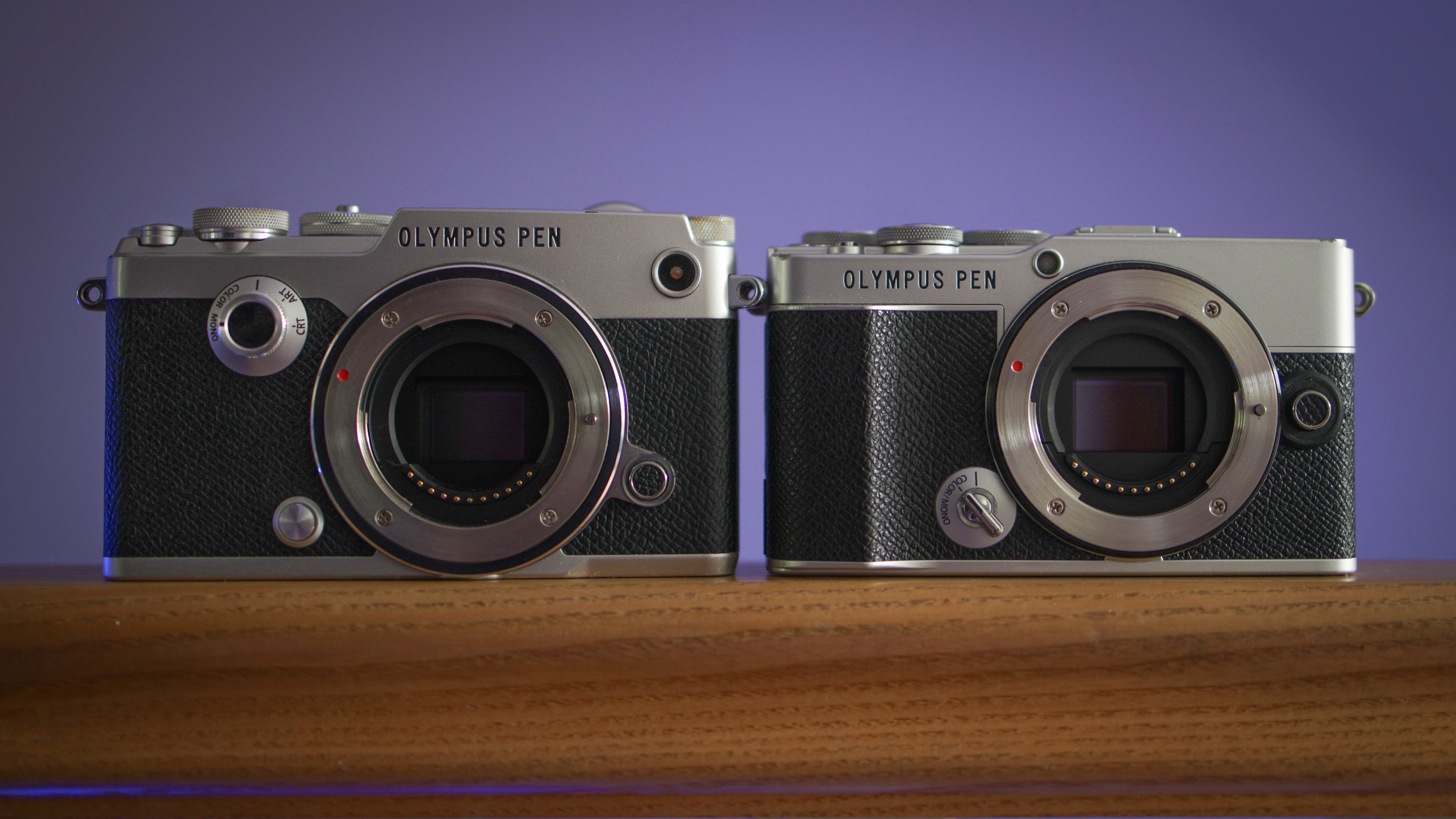 The Olympus PEN E-P7 side by side next to an Olympus PEN-F on a wooden surface against a purple background