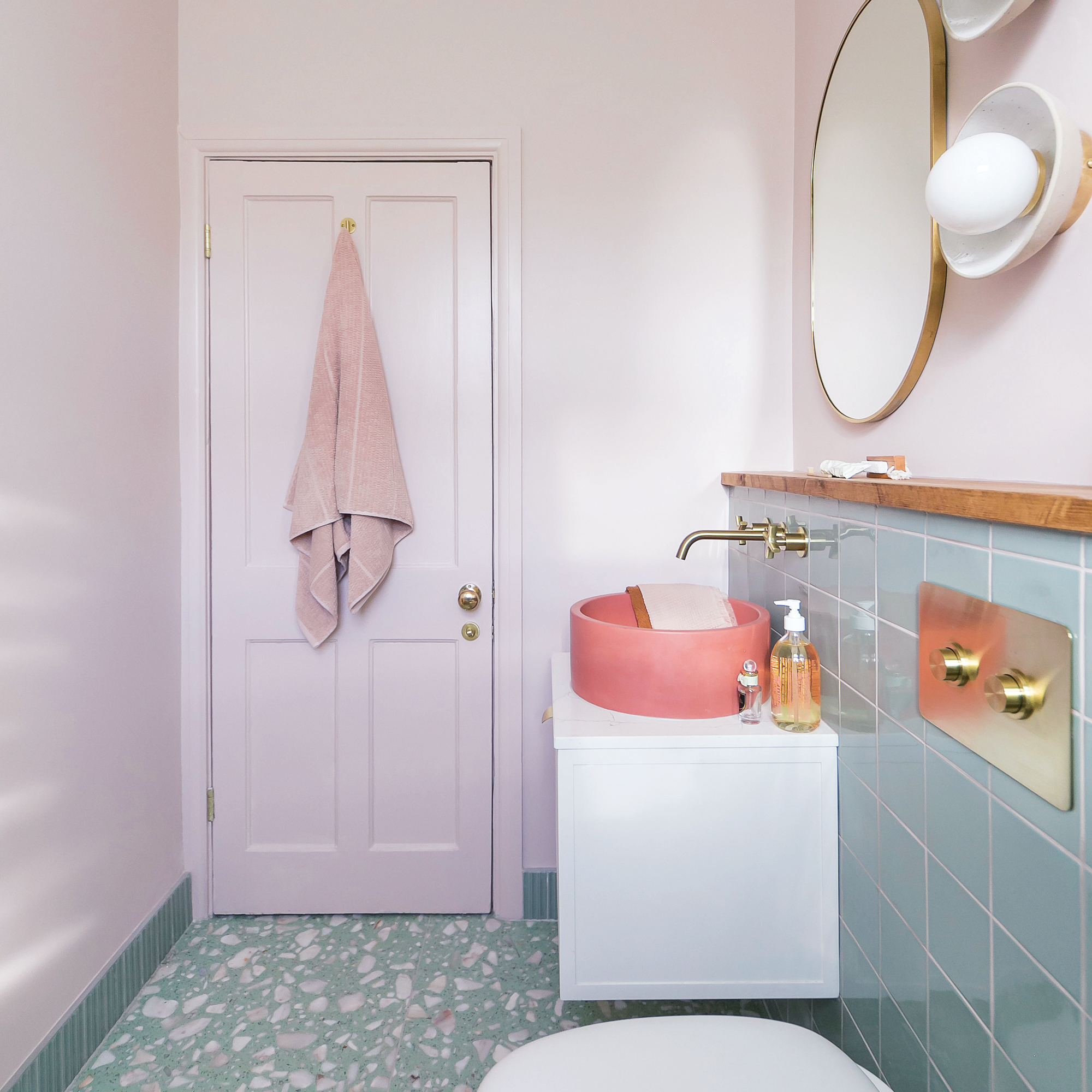 a small bathroom with pink painted walls, green wall tile, bright coral modern bowl basin and green terrazzo flooring