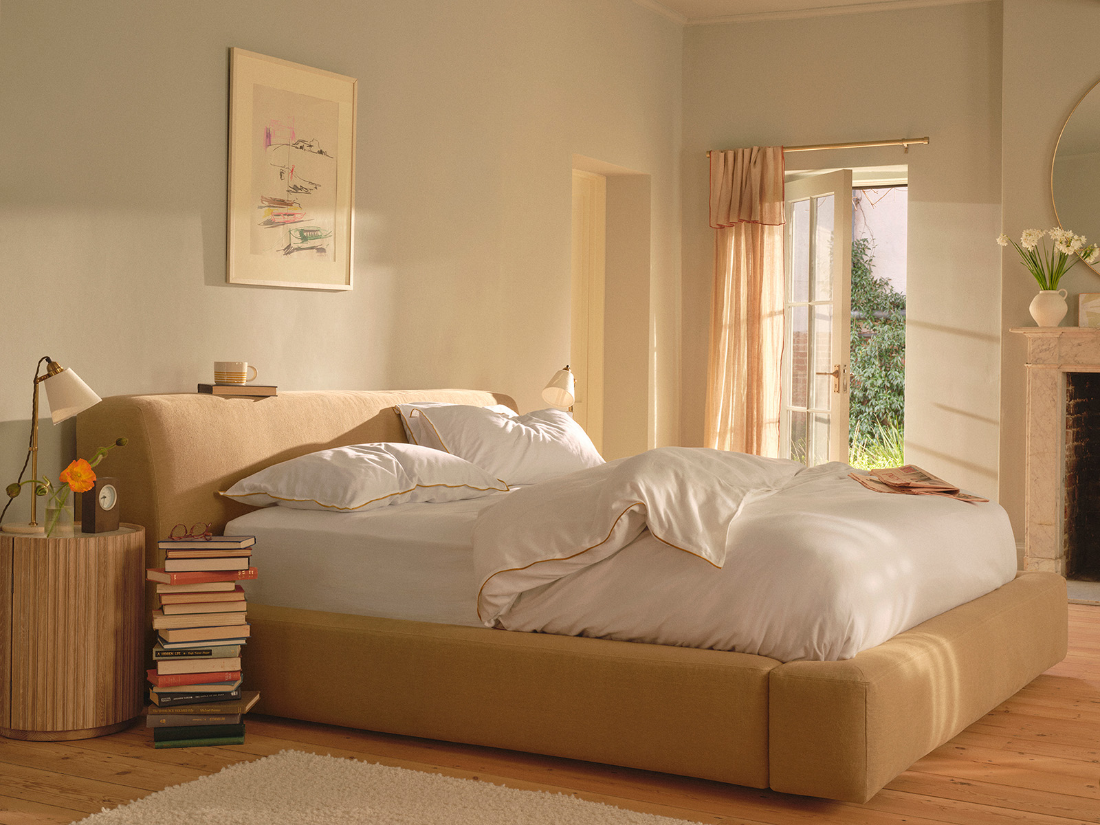 beige bed in a gray bedroom with stacks of books next to a wood nightstand