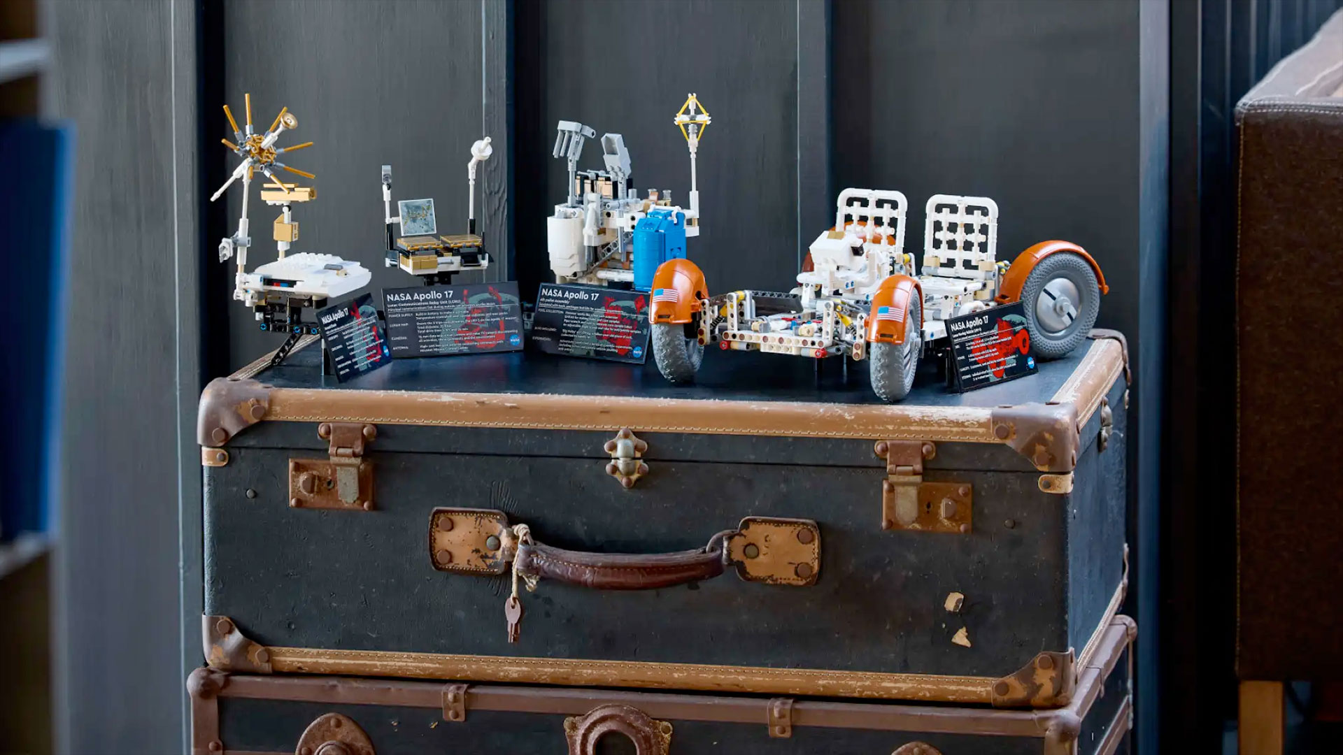 a white lego moon rover and several other small builds sit on top of a brown and black trunk against a gray wall.