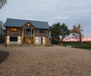 A timber clad two storey house with a wide gravel driveway leading up to it at sunset