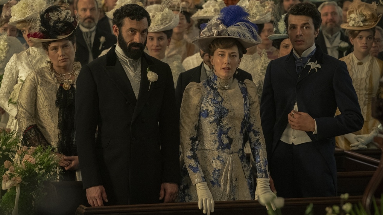 A lavishly dressed crowd gather at an indoor event, fronted by three concerned-looking members of the Russell family: dad George (Morgan Spector), mum Bertha (Carrie Coon), and their son Larry (Harry Richardson), in The Gilded Age Season 3.
