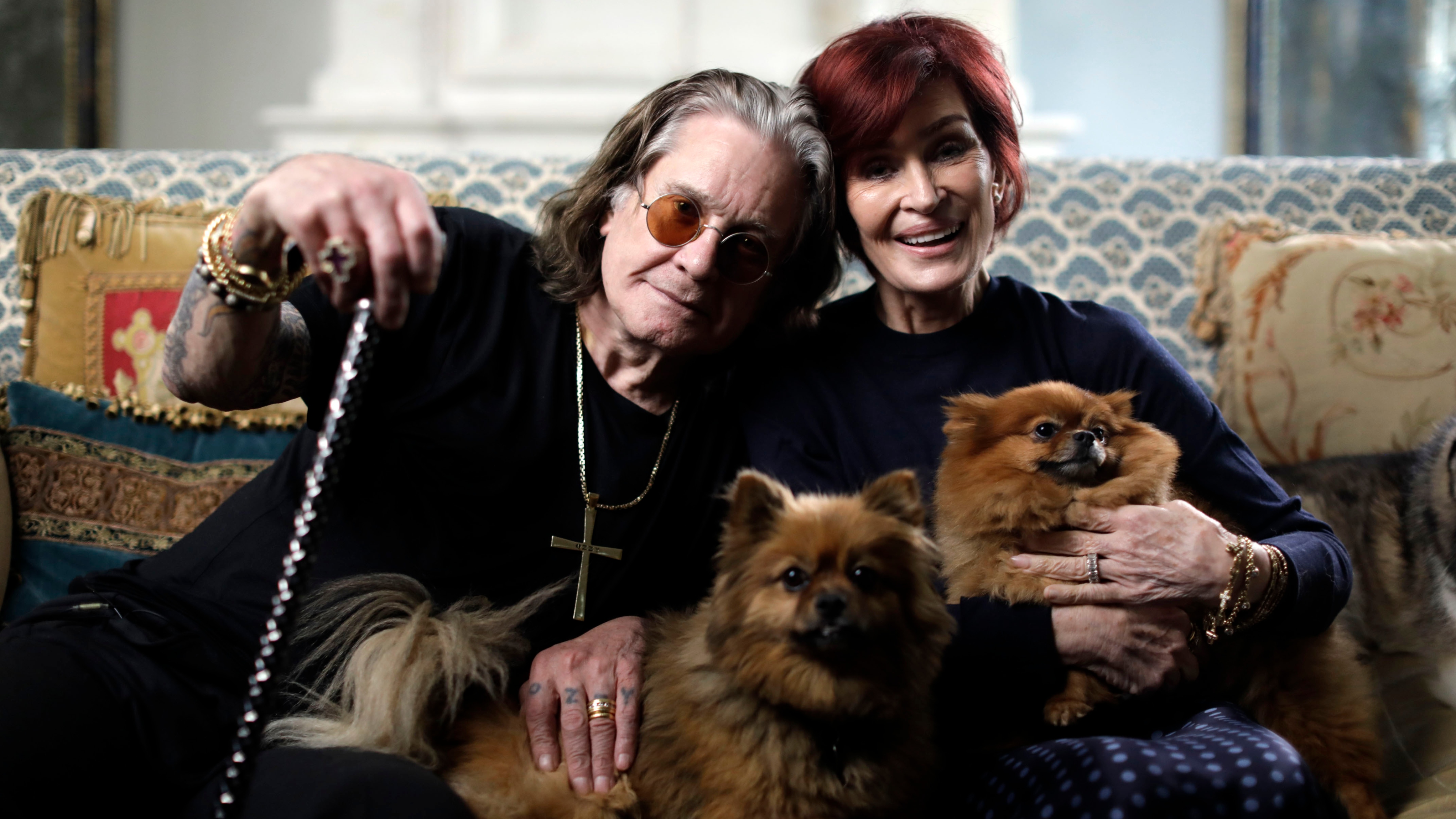 Ozzy Osbourne and Sharon Osbourne sitting on a sofa with their dogs on their laps and smiling for the camera