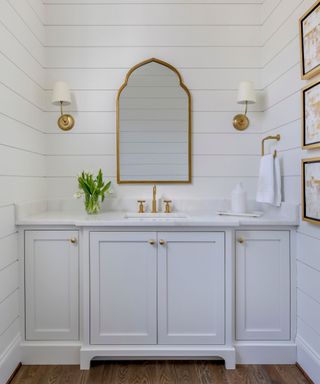 bathroom with white panelled walls, white cabinets and gold wall scones and mirror