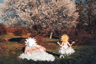 Two naked men sitting in the countryside on fabricated clouds with cut-out pictures of the sun covering their faces