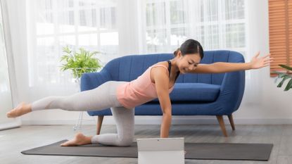 east asian woman wearing pink vest grey leggings blue sofa behind her doing bird dog exercise