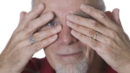 An older man peeks through the fingers of one hand while having both hands over his eyes.
