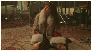 Riley Keough (Daisy Jones) sat on the floor wearing a colorful fur-trimmed jacket in Amazon Prime's Daisy Jones & The Six