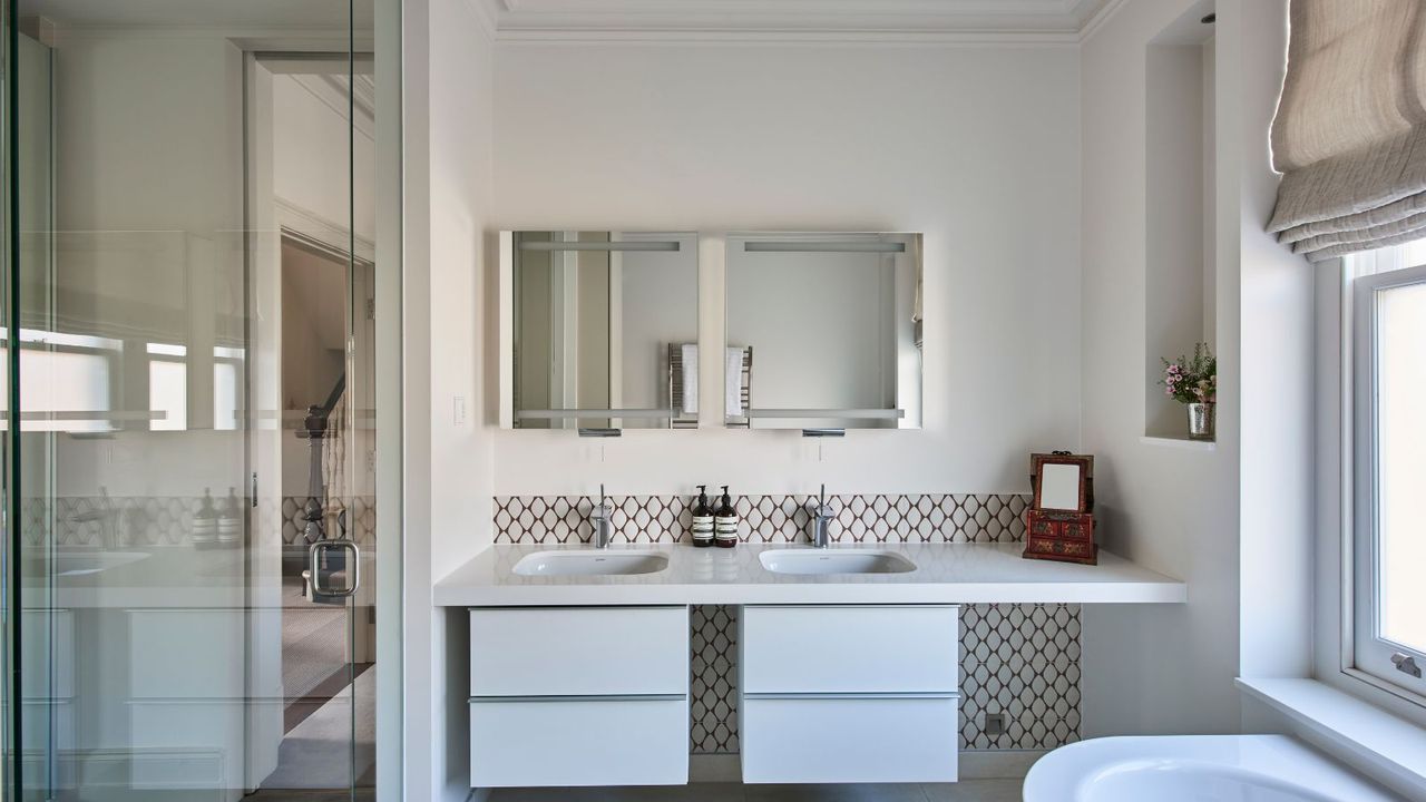 A bright bathroom with white basins with tiled backsplash, mirrored cabinets and a glass shower screen opposite a bright window with a neutral blind above a white porcelain bathtub