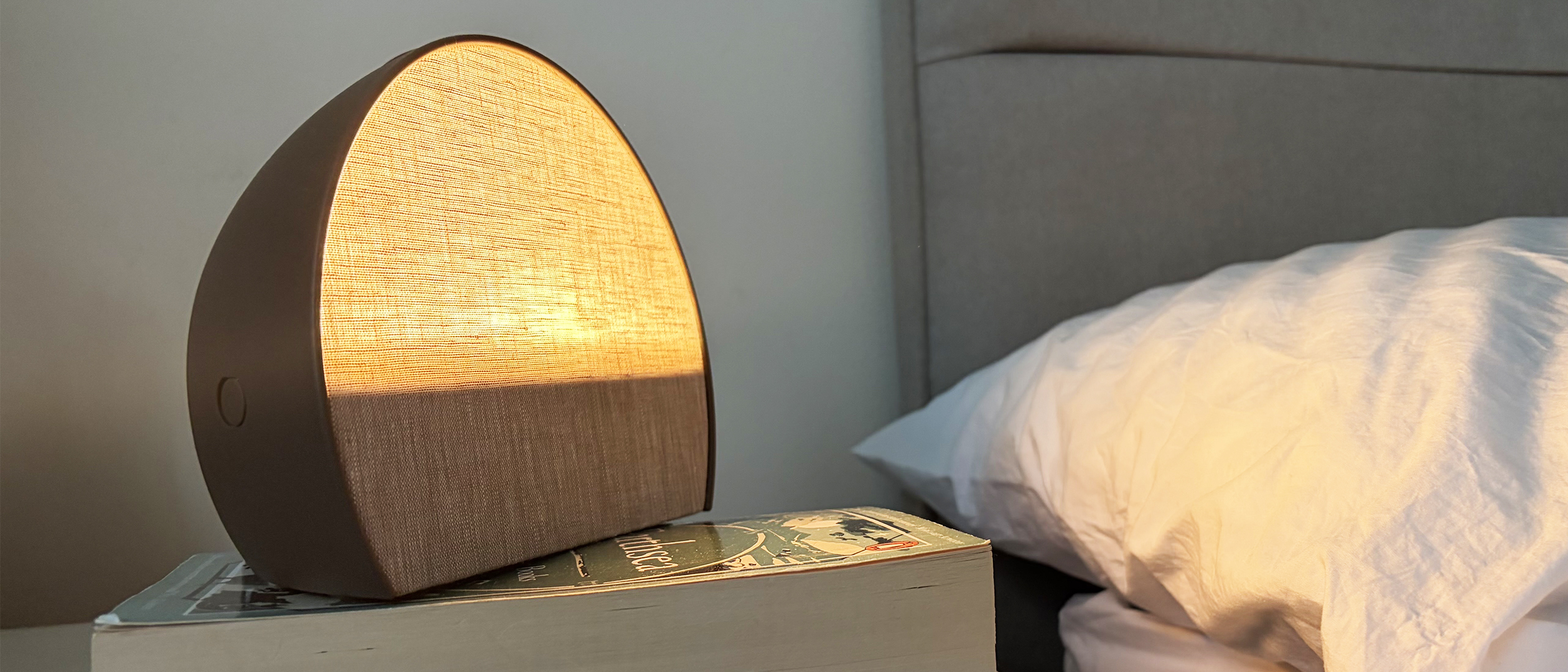 A close up of the Hatch Sleep Clock sunrise alarm clock and sleep tracker lit up on a nightstand next to a pillow