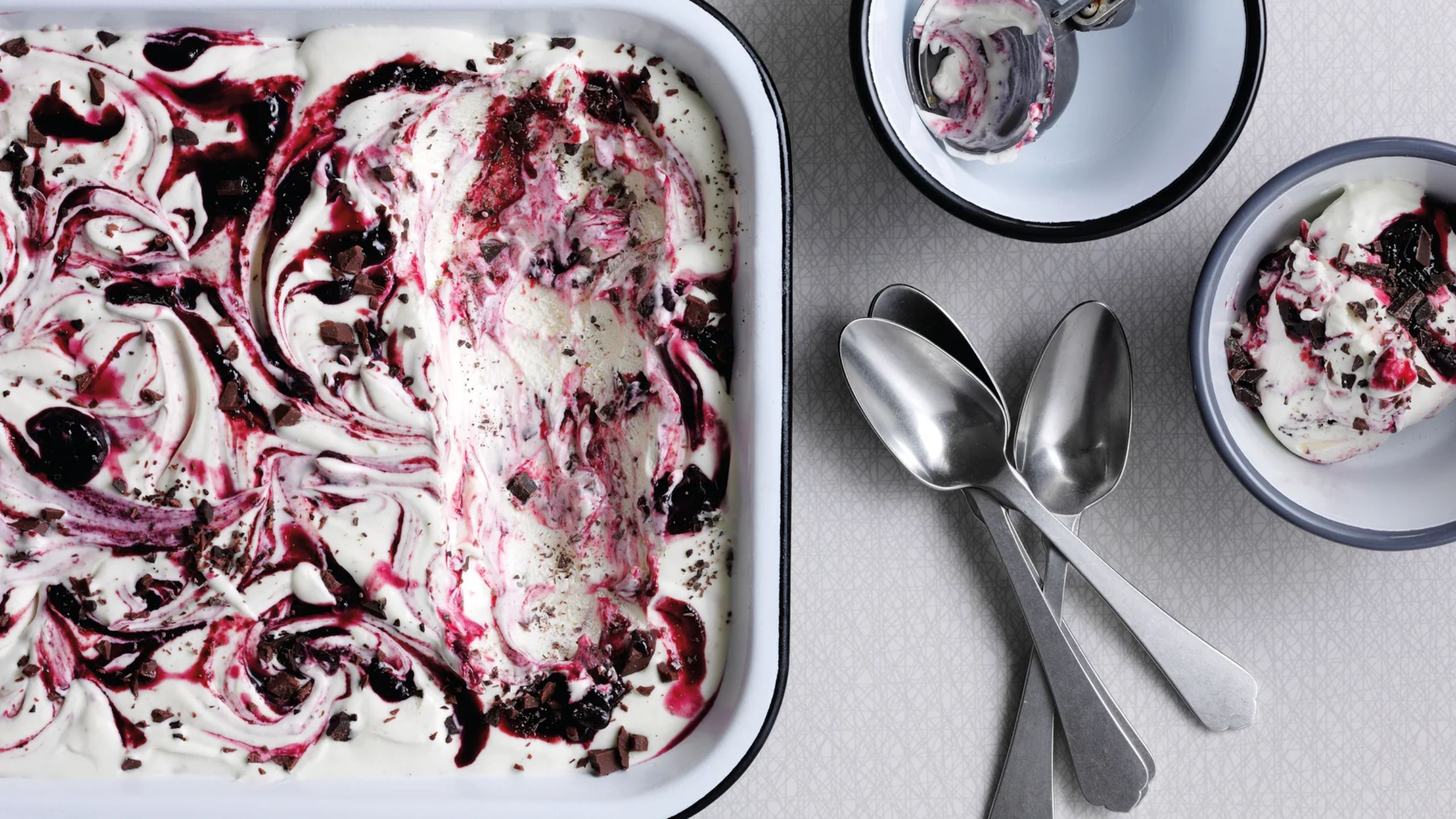 A white enamel dish with a black rim, with swirled ice cream inside, beside two bowls with scoops, and three teaspoons. 
