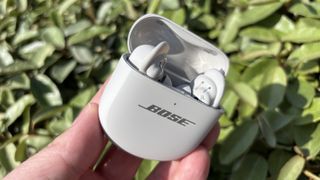 Bose QuietComfort Ultra Earbuds (2nd Gen) being held in a hand in their case with a green bush as a backdrop