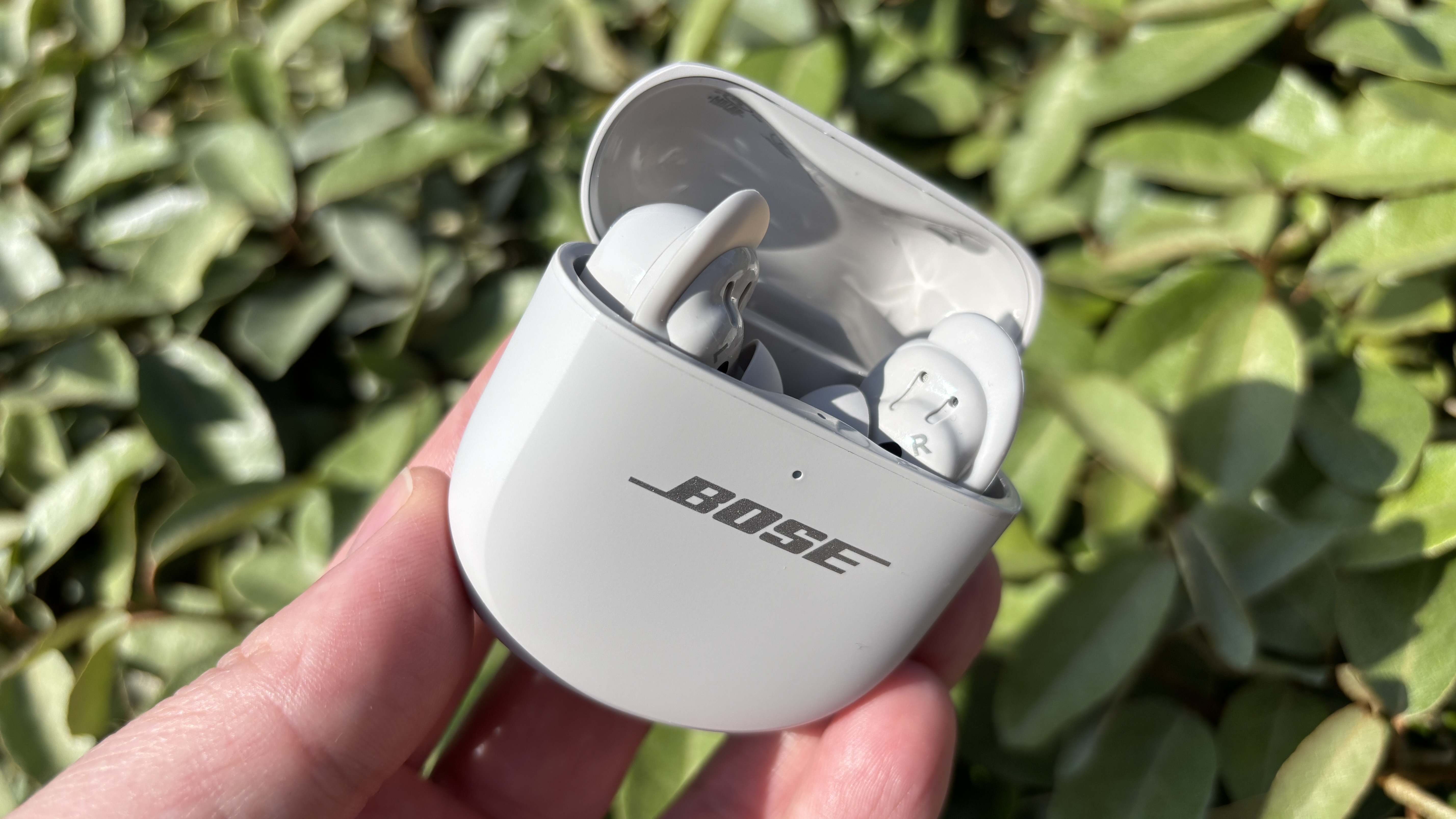 Bose QuietComfort Ultra Earbuds (2nd Gen) being held in a hand in their case with a green bush as a backdrop