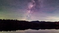 starry night above a lake. the milky way stretches up from behind silhouetted trees to fill the center portion of the sky in the image.