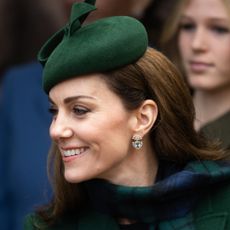 Kate Middleton wearing a blue and green plaid scarf and green hat looking to the left and smiling