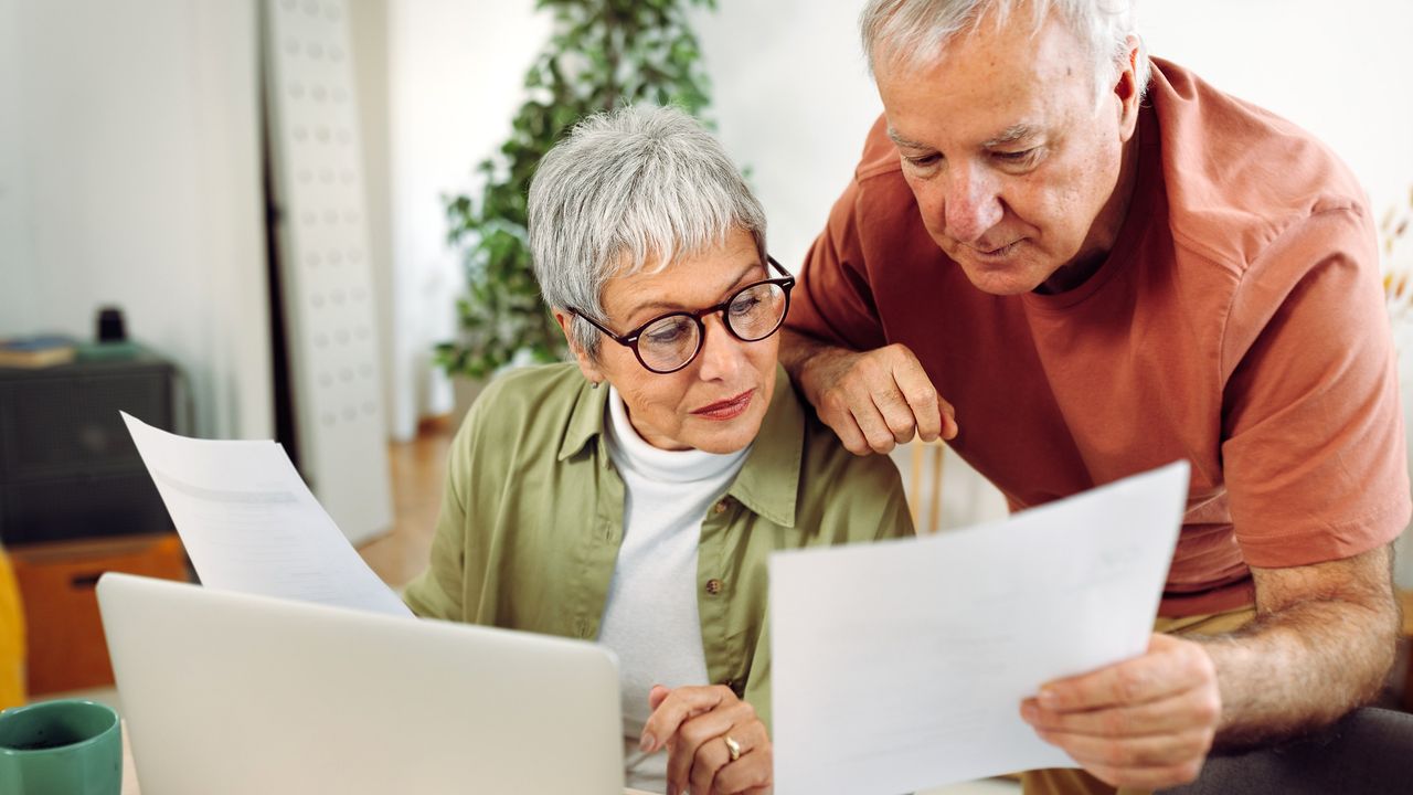 An older couple look over multiple forms together at their kitchen table.