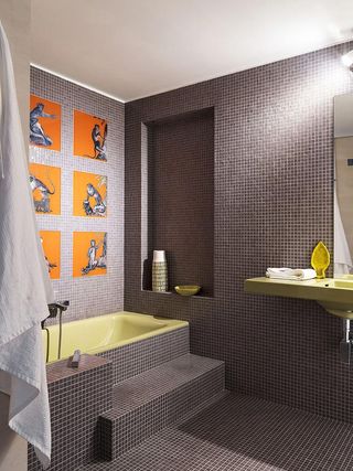 Interior view of a bathroom at House of Birds in Portugal featuring small dark grey tiled walls and floor, a white ceiling and a pale yellow sink and bath. There are steps leading up to the bath, an alcove with decorative pieces and six orange decorative squares with animals on the wall