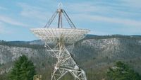 a radio antenna dish points toward the blue sky above a mountain ridge