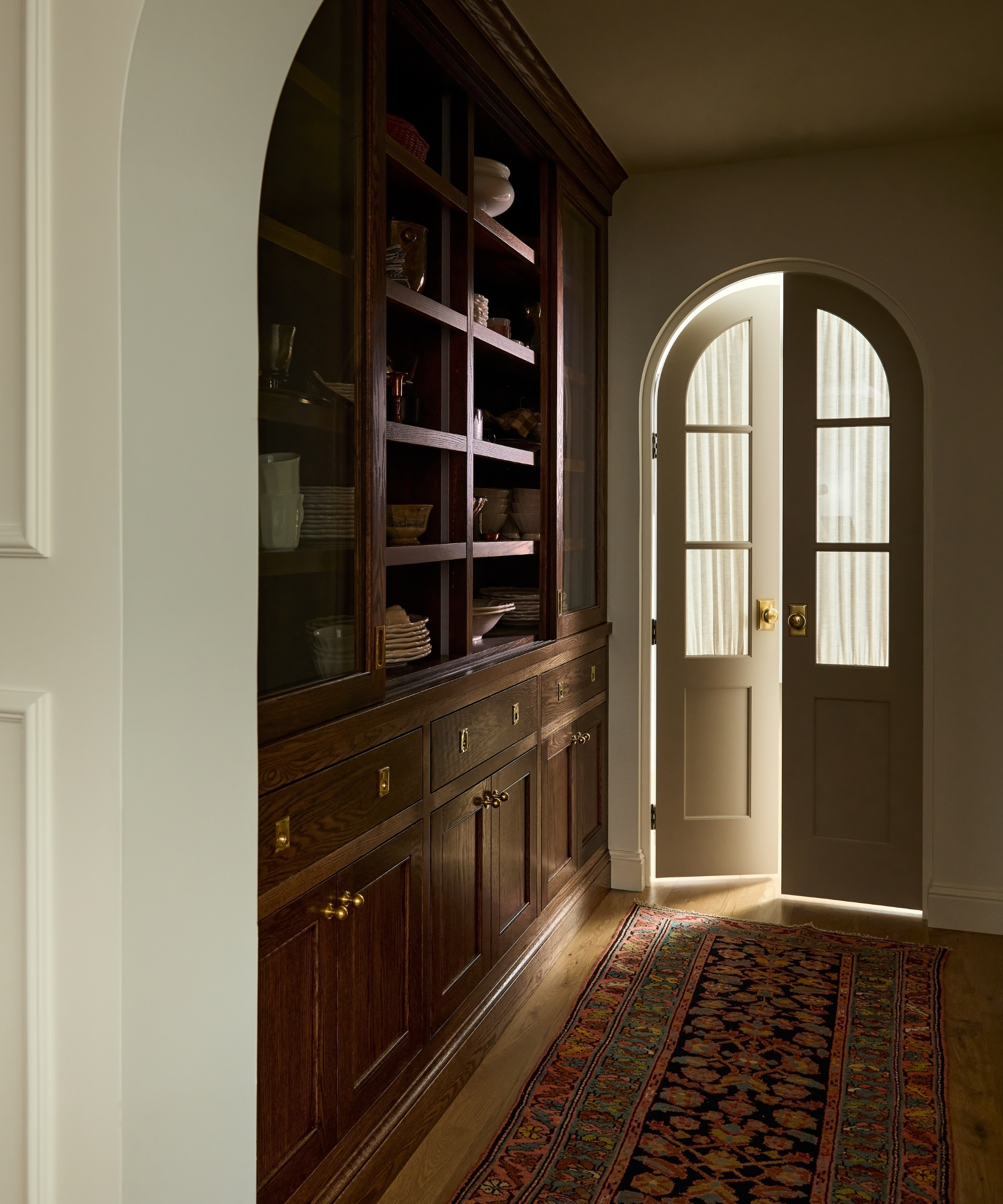 hallway of a lake house with a large dark wood pantry wall storage and an arched glass door with a vintage red toned runner rug
