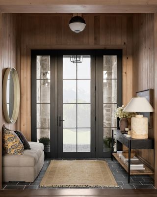 A light yellow rug in a modern entryway. The walls are wood panelling and there is a white fabric bench seat with a circle mirror above it on the left, and a black console table with a light yellow lamp on it on the right