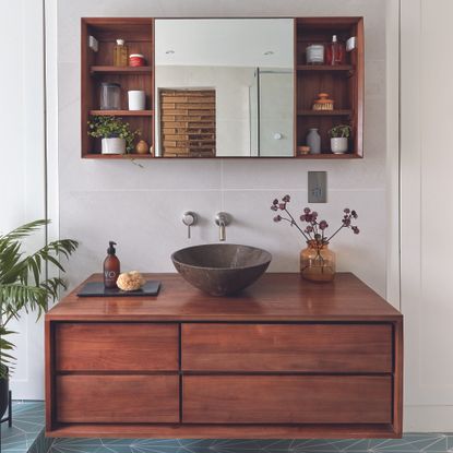 Dark wood bathroom storage unit in bathroom with mirrored cabinet.