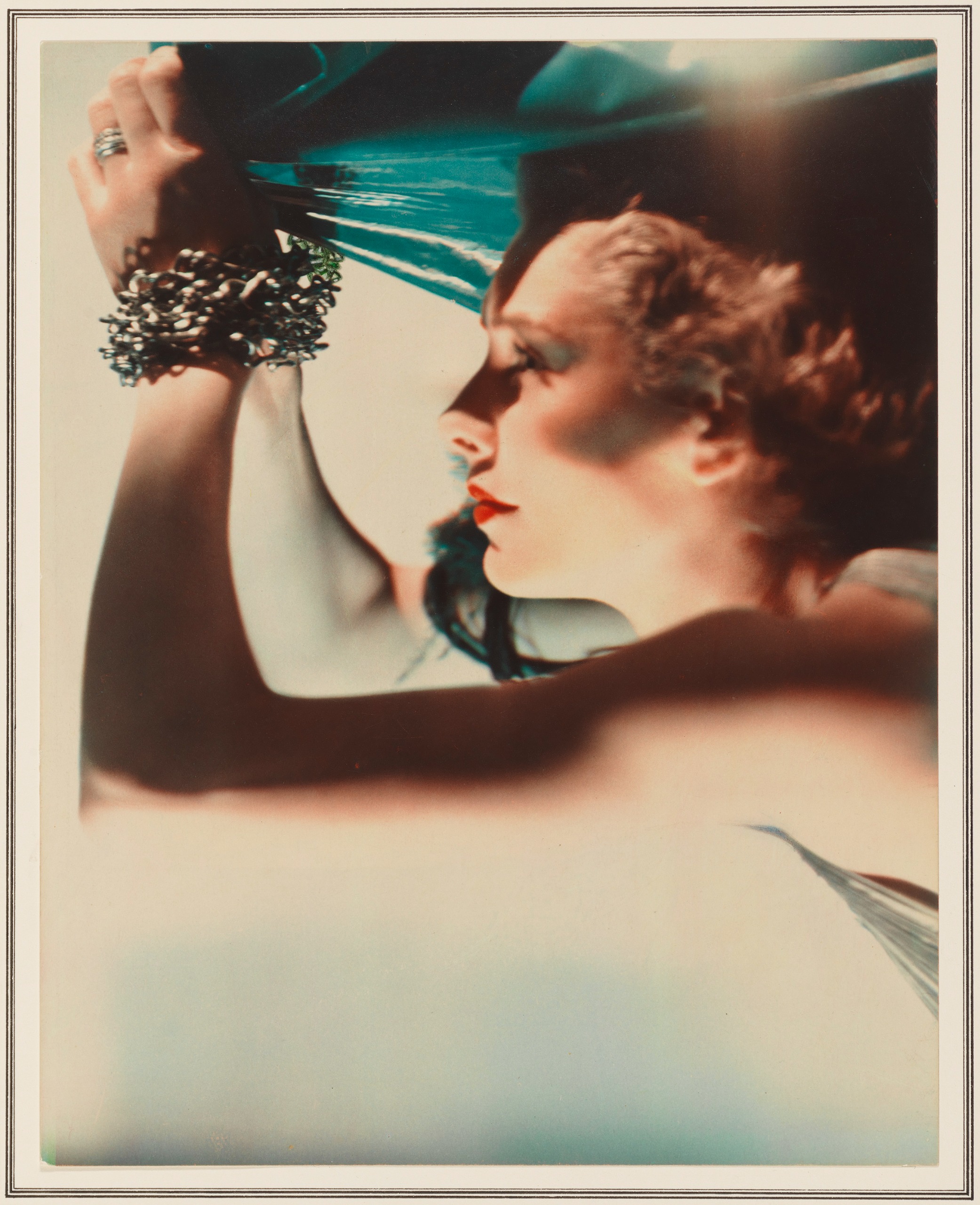 A color portrait of a woman in profile with curled hair and red lipstick, one arm raised and wrapped in ornate bracelets against a teal fabric backdrop.