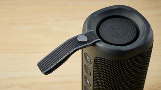 a closeup picture of the carry strap of a black oblong-shaped bluetooth speaker with mesh covering and a carry strap photographed on a beige table in front of a navy wall