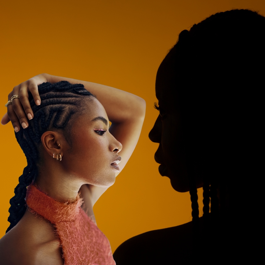an orange background with the shadow of a woman with braids in her hair facing a black woman in a pink top with braids in her hair