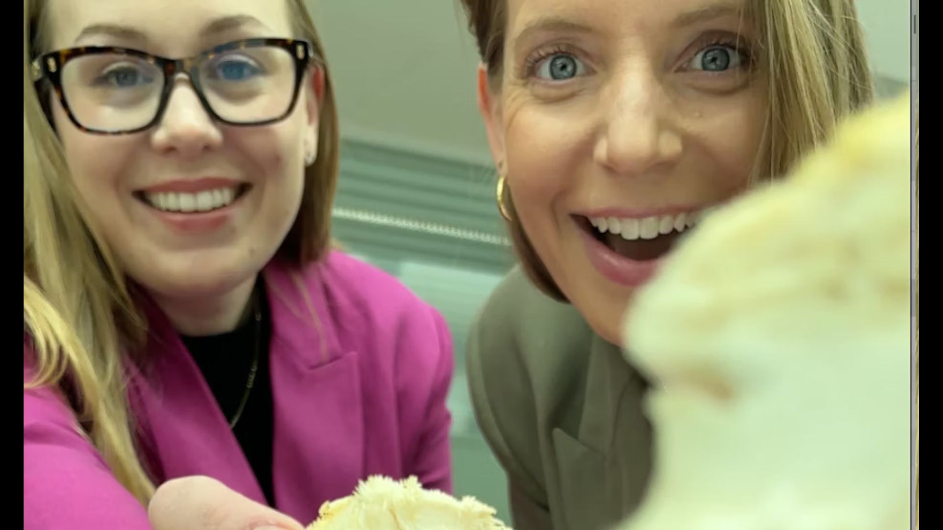 Two women hold up spongy white materials to the camera, one woman wears glasses and a pink blazer and the other has a gold hoop earring