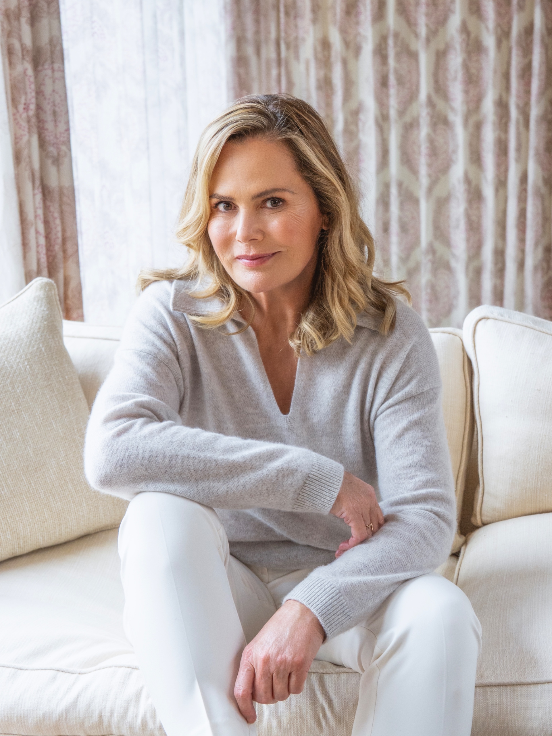 A blonde woman sitting on a cream sofa with one arm resting on her knee, wearing a grey jumper and white jeans