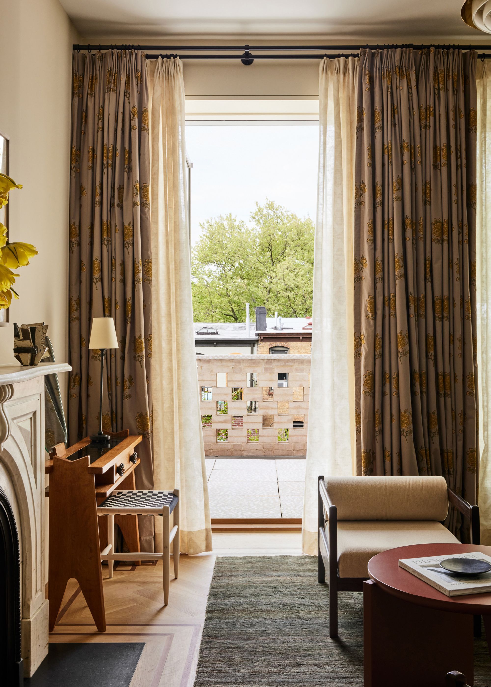 A living room with a study and stool, a stone fireplace, a sofa chair, a round coffee table, a textured rug, patterned curtains and a balcony