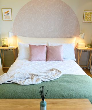 Bed with white sheets, pink throw cushions and textured green throw with calm graphic pink painted half circle above headboard and white wall sconces either side
