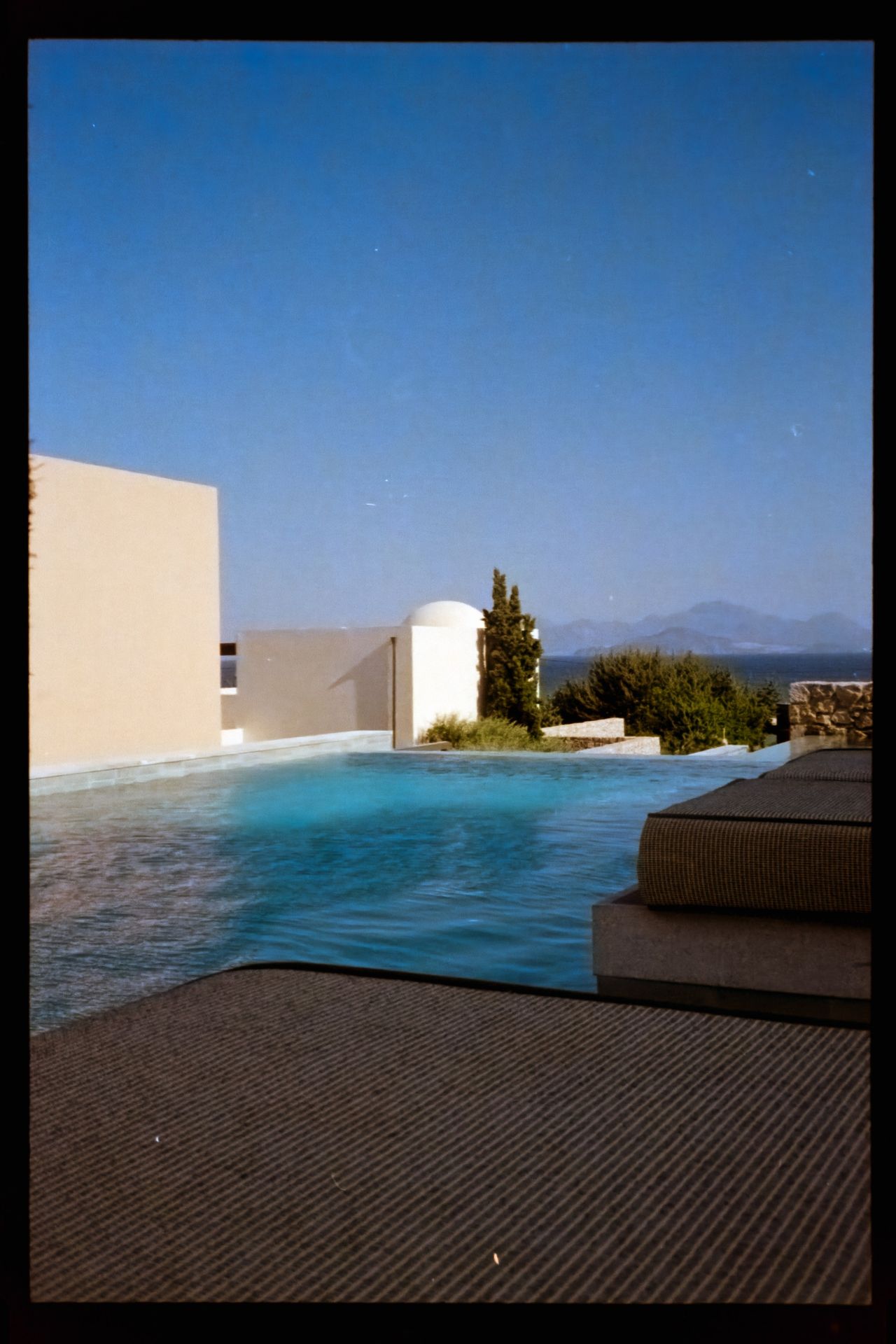Scanned film photograph of a modern outdoor swimming pool overlooking the sea, framed by white architecture and a clear blue sky.