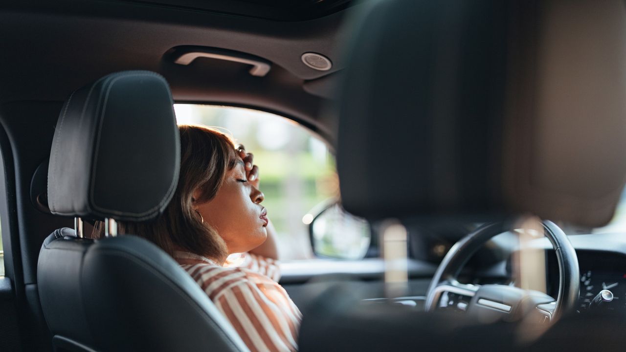 A woman sits in the driver&#039;s seat of her car. She has a hand to her head, her eyes are closed and she is breathing deeply.