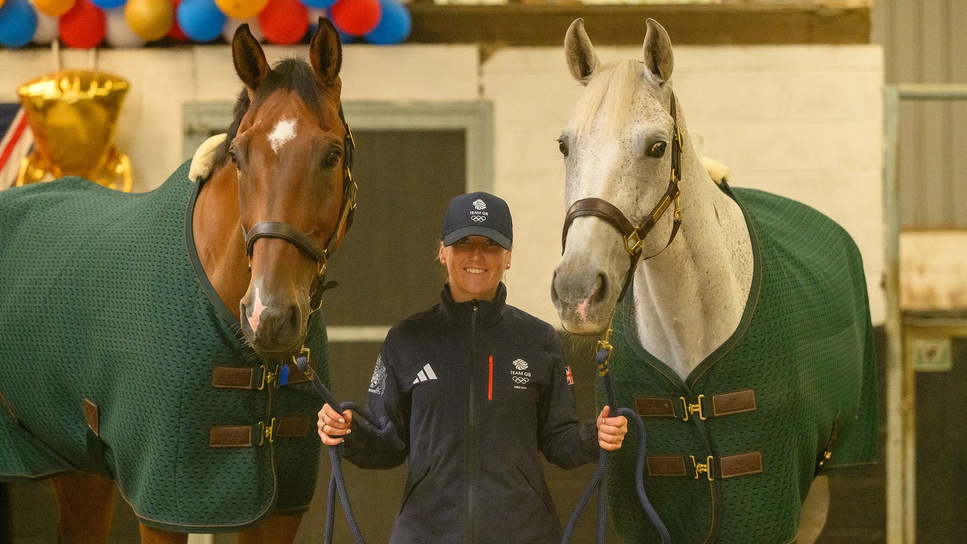 Laura Collett, a rider wearing a casual British team jacket and baseball cap, holds bay horse London 52 and grey horse Hester