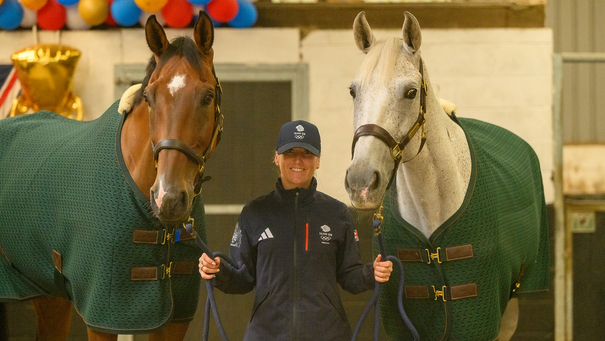 Laura Collett, a rider wearing a casual British team jacket and baseball cap, holds bay horse London 52 and grey horse Hester