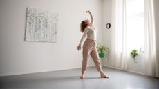 Woman doing ballet-inspired workout at home in open studio space, arms in the air and head back
