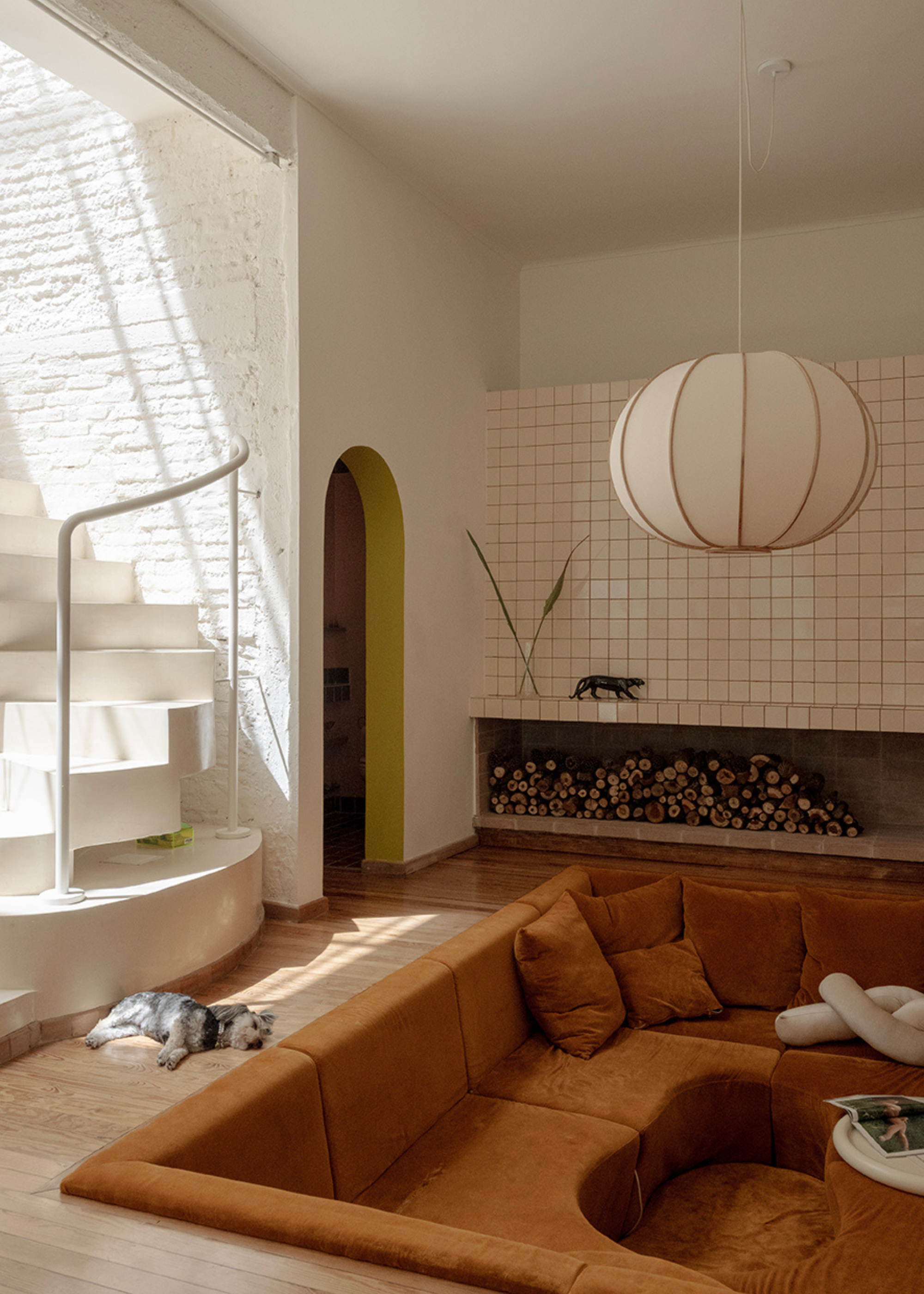 A living room with a tiled accent wall, a nook with logs, an amber conversation pit, and a sleeping dog beside a curved white staircase
