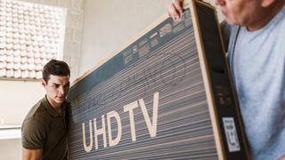 Two people carry a TV box through a room. The box is labeled "UHD TV."