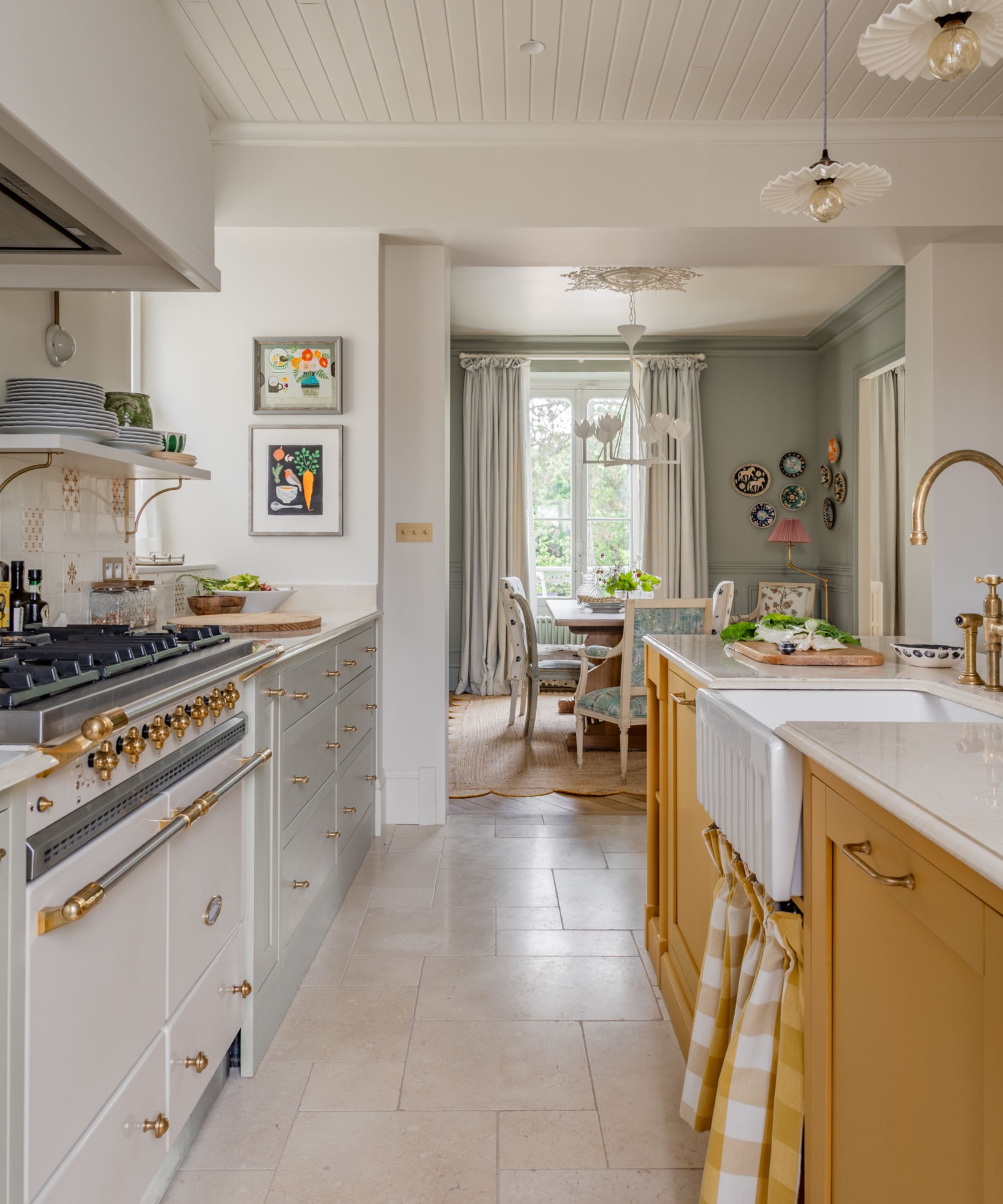A perspective shot looking down a light stone tile floor from the kitchen into a cozy dining room. A white farmhouse sink is in the foreground, and a large white range with brass hardware sits to the left