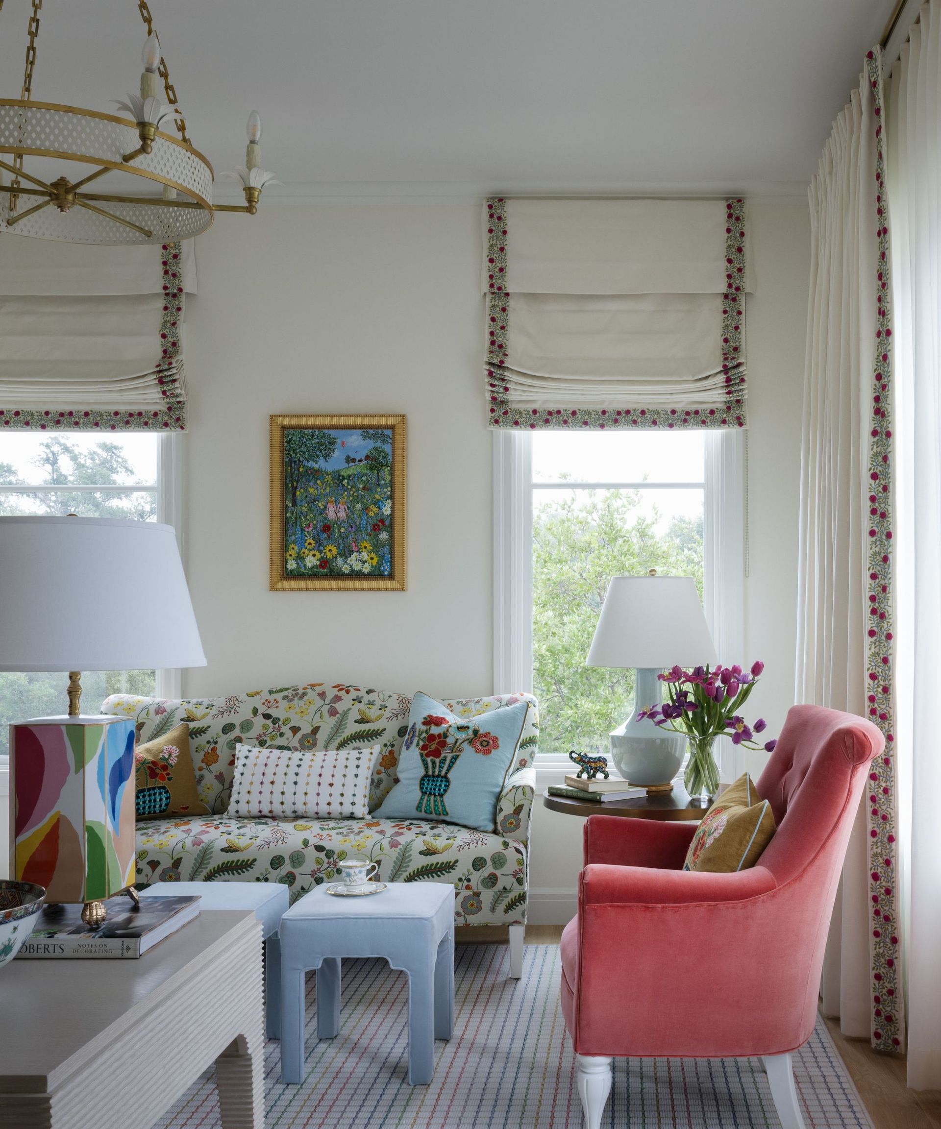 colorful living room with white walls and a floral couch, blue ottomans, pink armchair and floral trim window treatments