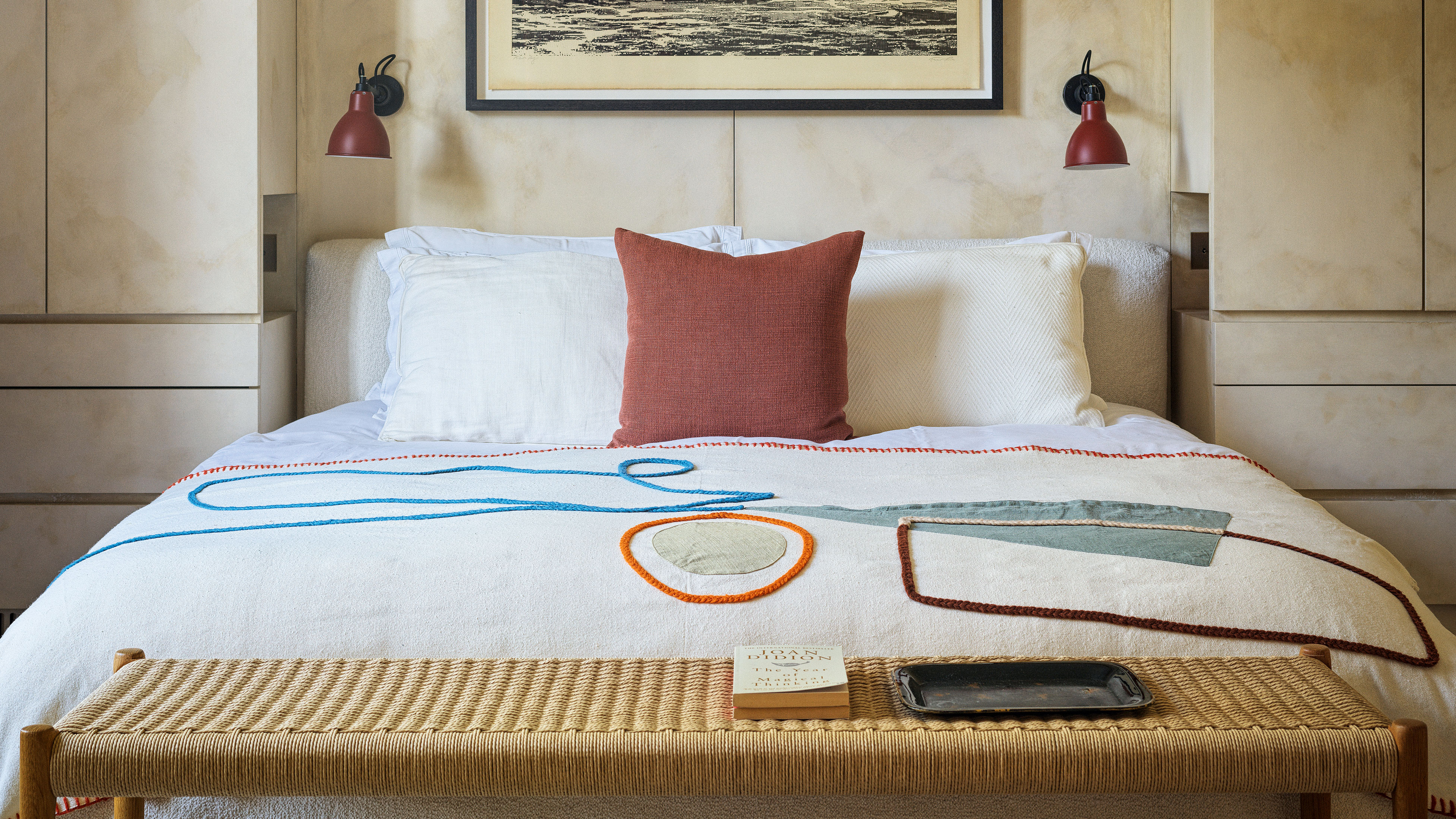 A brightly lit, modern bedroom featuring a bed with a white textured duvet decorated with abstract, colorful embroidered shapes in blue, orange, and green. At the foot of the bed sits a woven light-wood bench holding a book and a black tray. The headboard is flanked by built-in light wood cabinets and two small red wall-mounted sconces. Above the bed hangs a large framed horizontal piece of art.