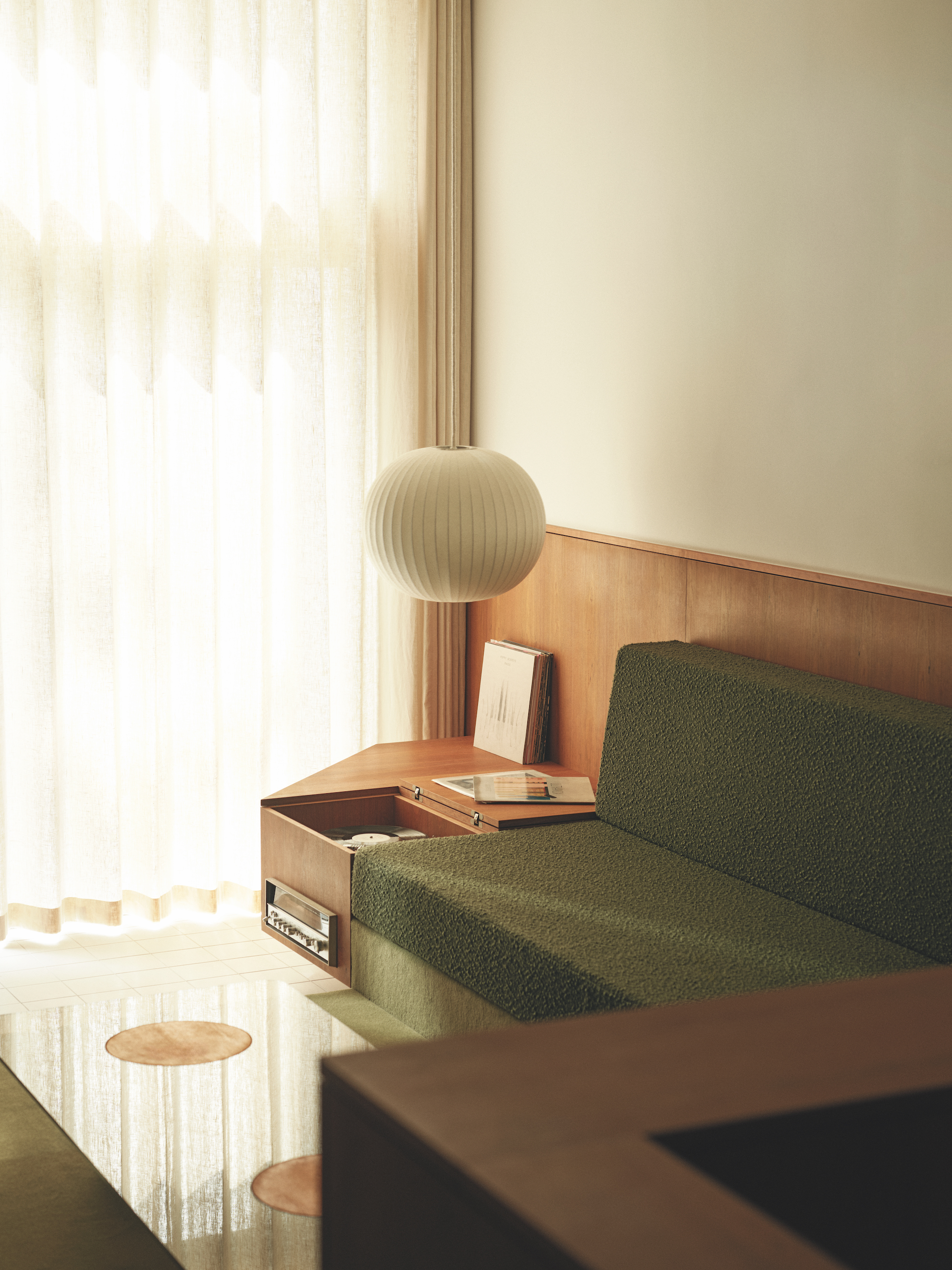 A view of the a built in sofa in a modern living area, the integrated bench is made from a dark wood with an olive green fabric for the sofa and a glass coffee table in front and cream curtains behind.