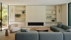 A bright living room with a blue sectional sofa, a white boucle armchair, wooden coffee tables, a wood side table, a white entertainment case and white shelves with vases and ceramics