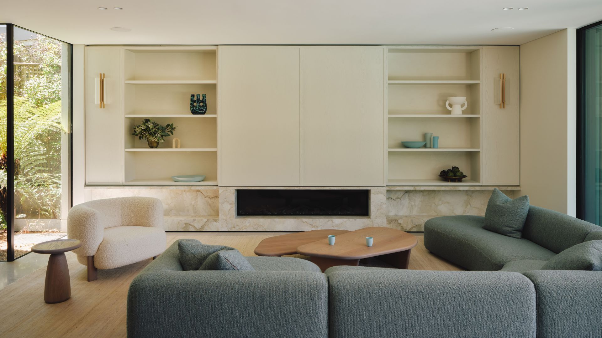 A bright living room with a blue sectional sofa, a white boucle armchair, wooden coffee tables, a wood side table, a white entertainment case and white shelves with vases and ceramics