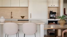 Image of a modern kitchen with cream cabinets and tan marble countertops. There are two blush pink barstools in front of the kitchen island and on the back counter there is a coffee station with an espresso maker.
