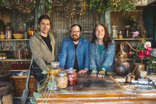 Handyman Tom Reade, Banjo and decorator Lisa McKenna pose, smiling, for a photo inside the apothecary. There are glass jars on the table in front of them filled with colourful ingredients, lots more on the shelves behind them, and herbs hanging from the ceiling