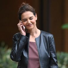 Katie Holmes walking by her apartment wearing a collarless motorcycle jacket with a burgundy pleated dress and slingback flats