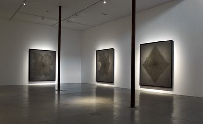 A gallery with large paintings on the walls with dark shapes on them and lights behind them.